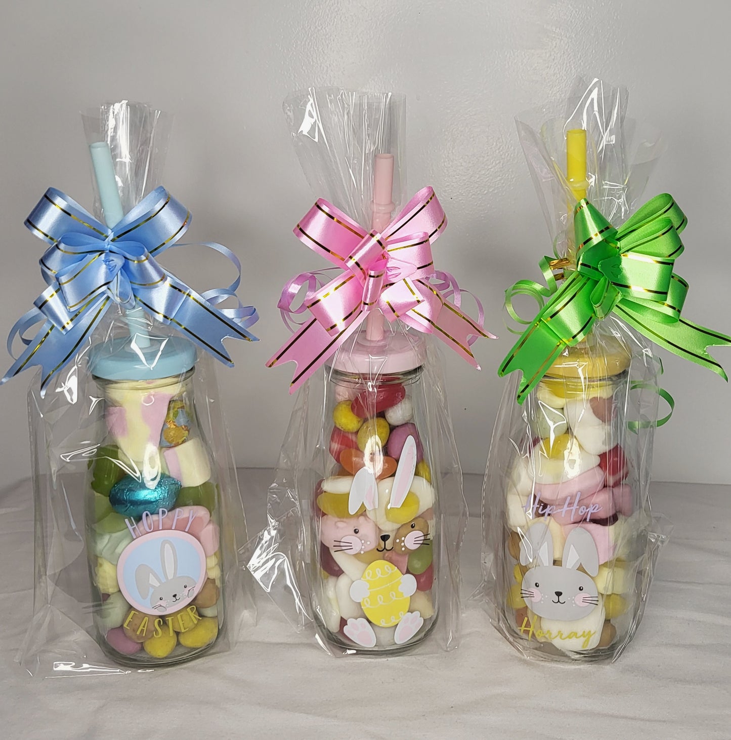 Three Easter-themed glass drink bottles with colorful decorative bows, filled with various candies and chocolates, presented in pink, blue, and yellow colors.