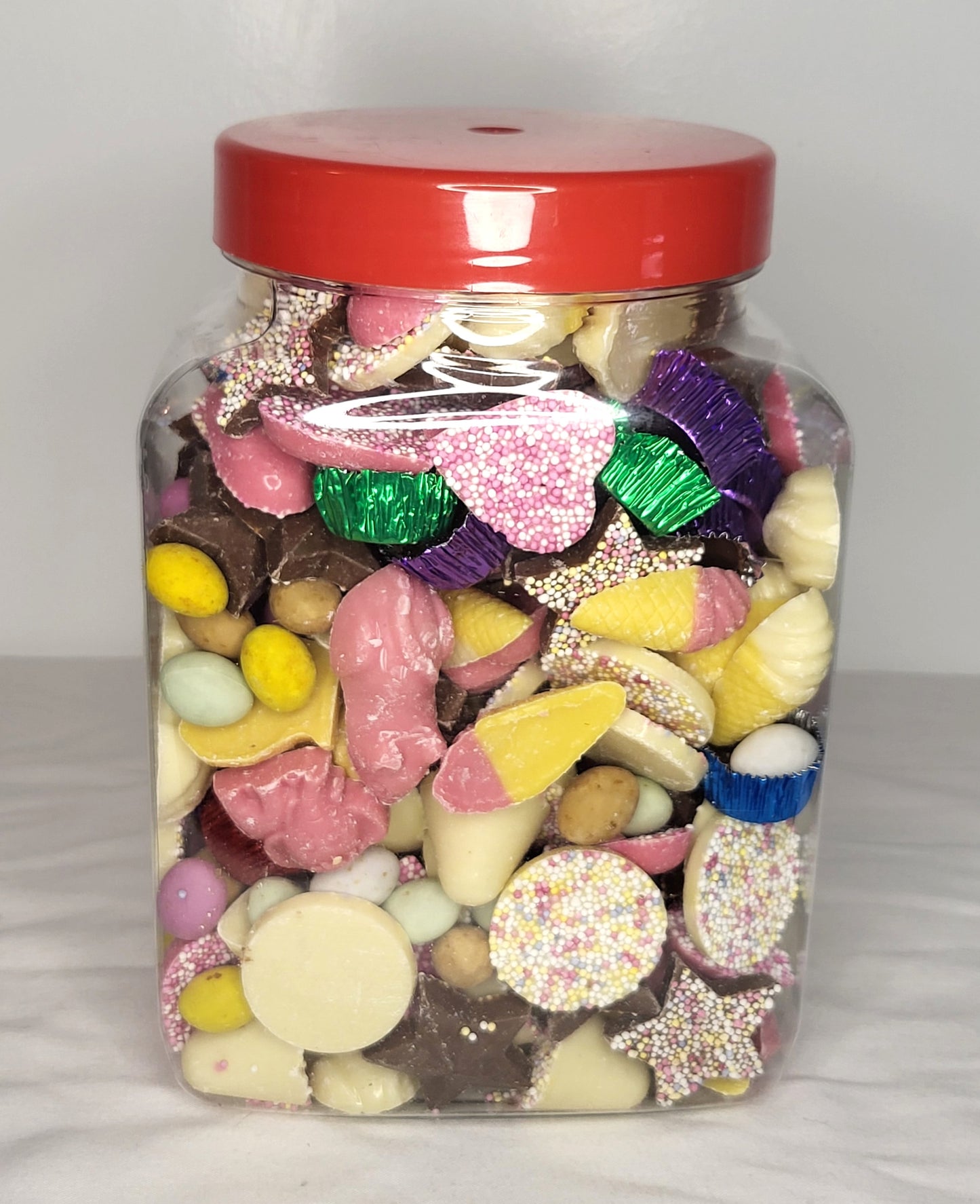 A medium-sized clear jar filled with a variety of chocolate candies and shapes, including hearts and eggs, sealed with a red lid.