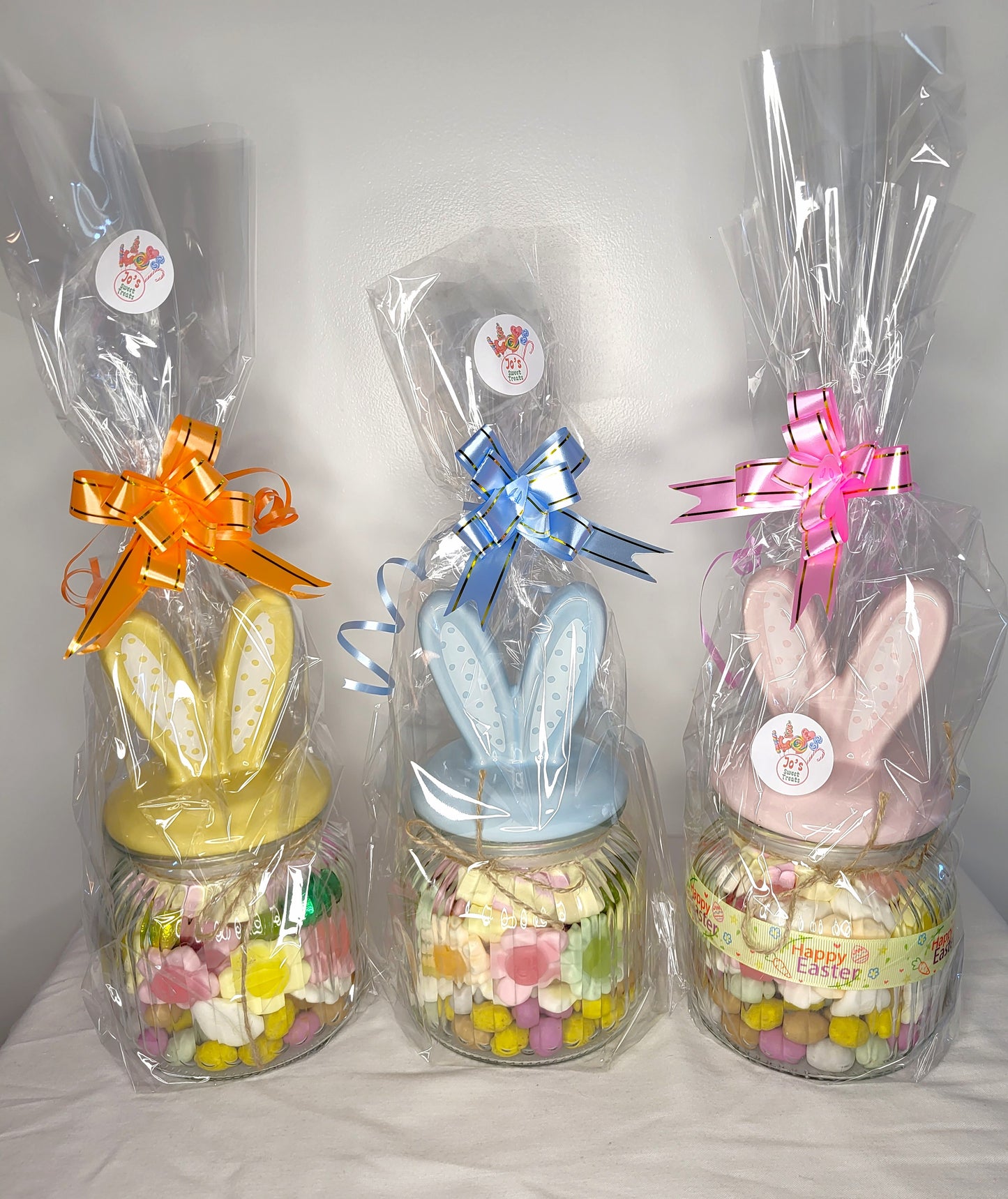 Three glass jars with bunny ears decoration, each containing different types of easter themed sweets, wrapped in plastic with orange, blue, and pink bows.